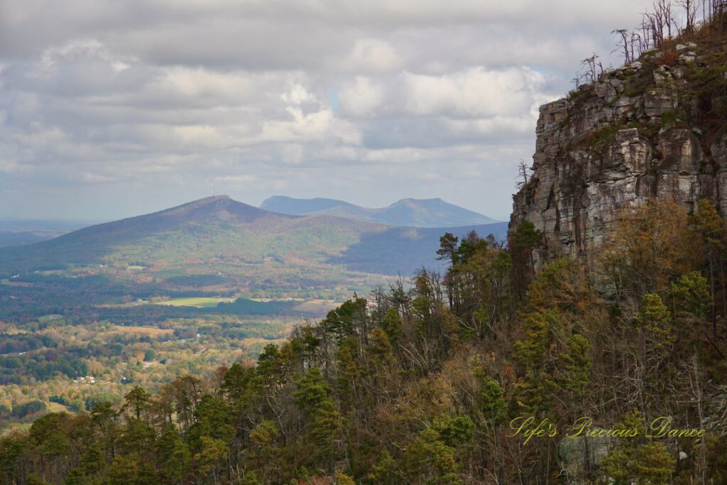 Close up of one side of Pilot Mountain on the right. In the distance on the left various mountain peaks can be seen. Passing clouds overhead.