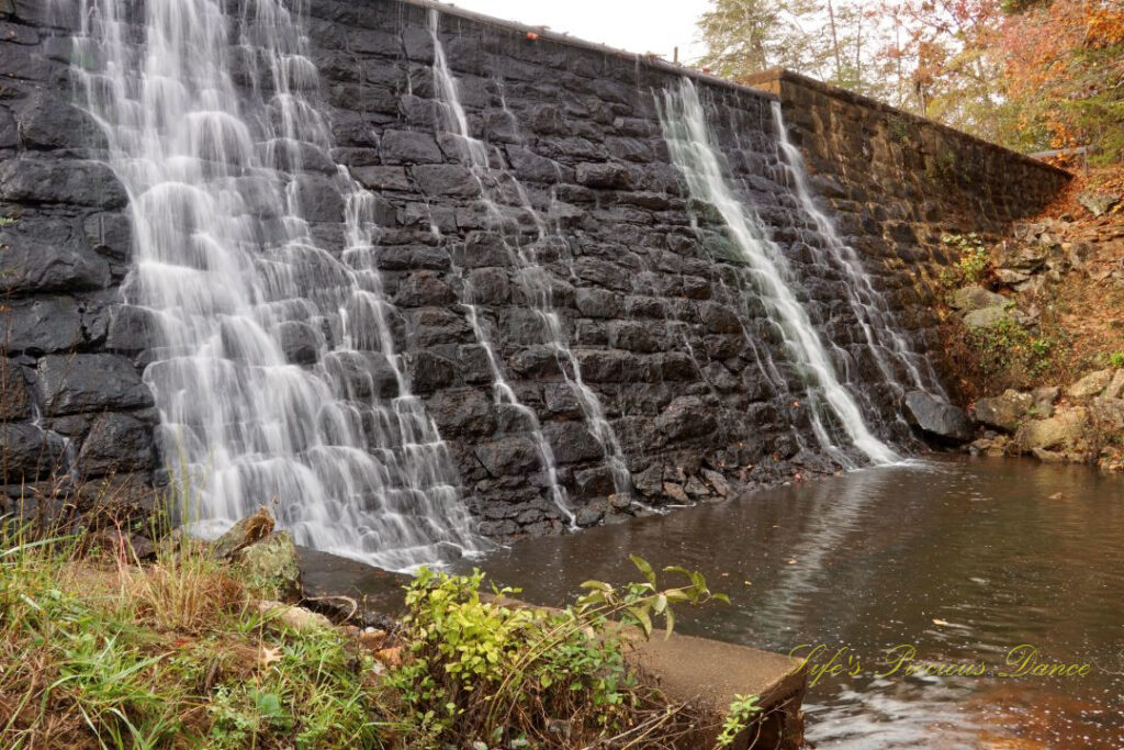 Mini waterfall spilling down a rockwall into the awaiting creek below. Colorful trees in the background.