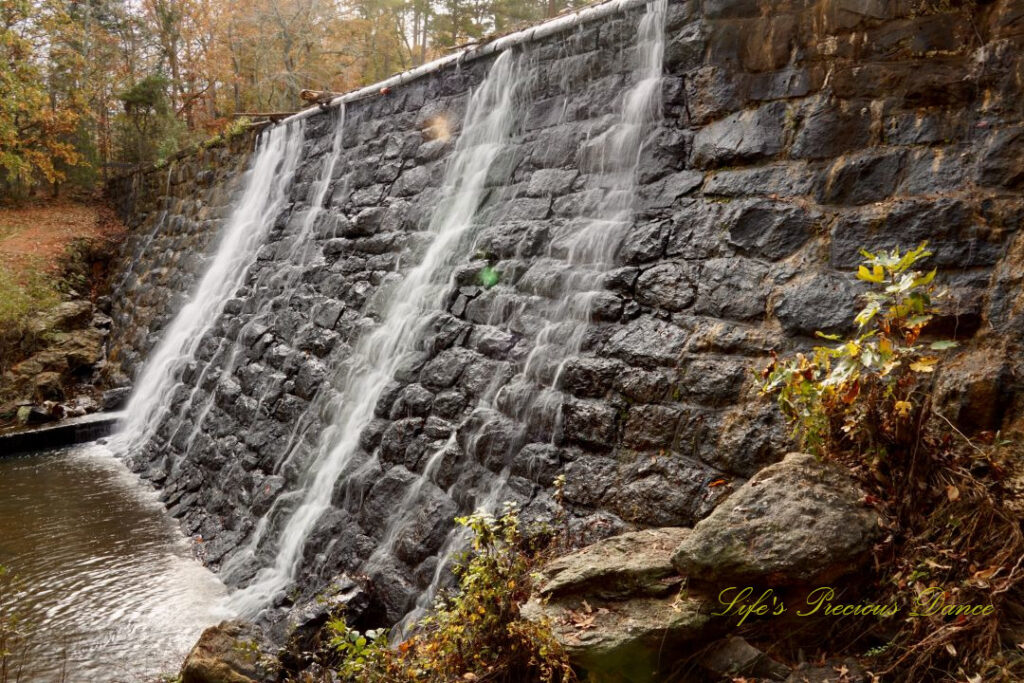 Side view of a mini waterfall spilling over a rockwall, into the awaiting creek below. Colorful trees in the background.