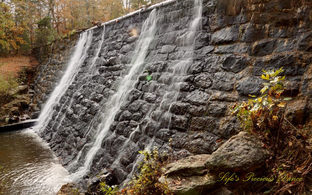 Side view of a mini waterfall spilling over a rockwall, into the awaiting creek below. Colorful trees in the background.